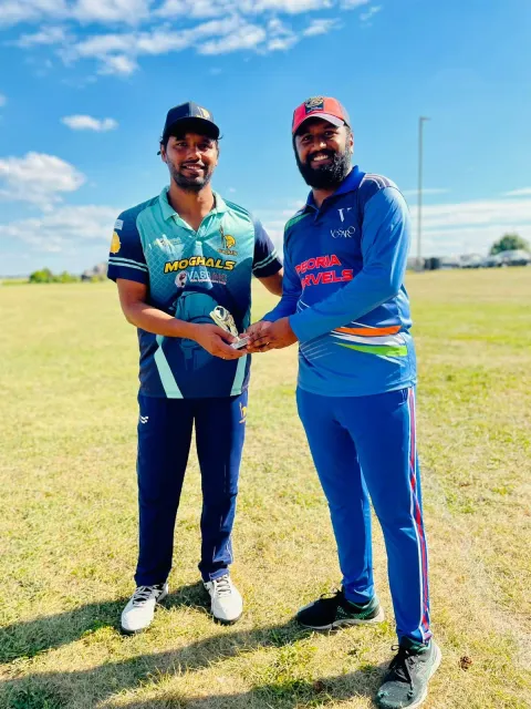 Two cricketers holding a trophy together on an outdoor cricket field.